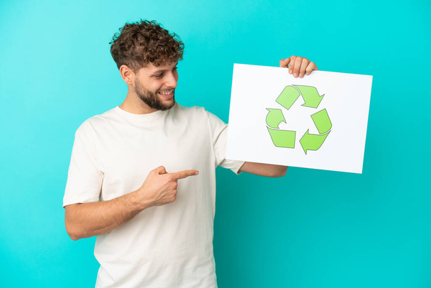 Junger gutaussehender kaukasischer Mann auf blauem Hintergrund, der ein Plakat mit Recycling-Symbol hält und darauf zeigt - Foto, Bild