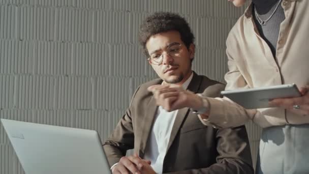 Středně naklápěcí snímek mladého muže ze Středního východu v brýlích a obleku sedícího na invalidním vozíku v kanceláři s laptopem a diskutujícího o projektové práci na obrazovce s bělošskou kolegyní s tabletem - Záběry, video