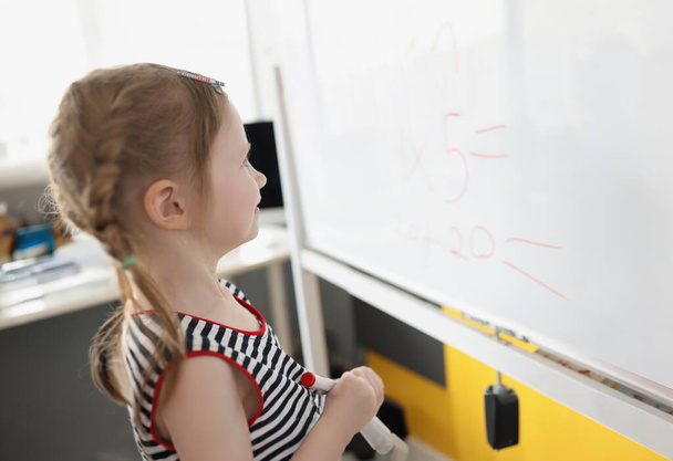 Портрет малыша, пишущего решения сумм на белой доске в школе, учебный процесс. Школьник думает, пока занимается математикой. Концепция - Фото, изображение