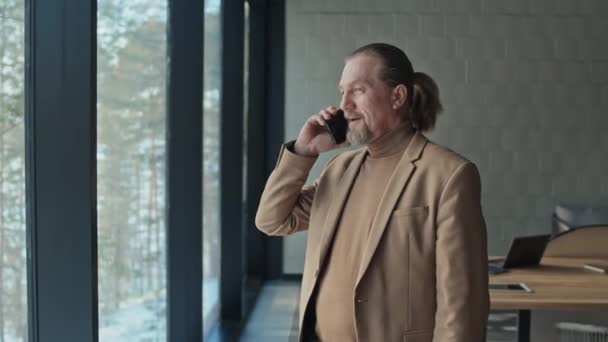 Mediana toma de maduro hombre de negocios caucásico en chaqueta beige y jersey de cuello alto hablando por teléfono móvil junto a ventana panorámica en sala de reuniones de hotel u oficina suburbana - Imágenes, Vídeo