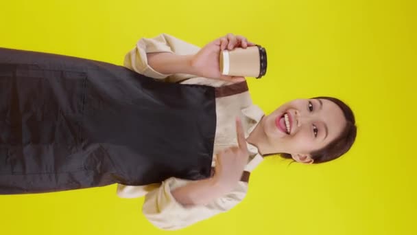 Portret jonge aziatische barista vrouw draagt schort met koffiebeker en duimen omhoog op gele achtergrond, serveerster of ondernemer met koffie met opgewonden, kleine onderneming of startup, ober van cafe. - Video