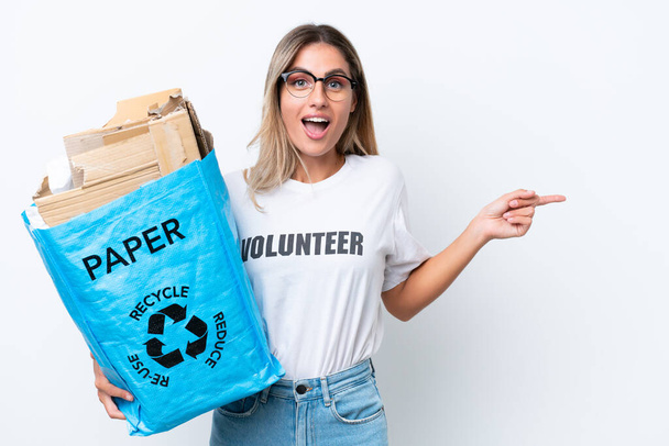 Joven guapa uruguaya sosteniendo una bolsa de reciclaje llena de papel para reciclar aislada sobre fondo de croma blanco sorprendida y señalando con el dedo hacia un lado - Foto, imagen