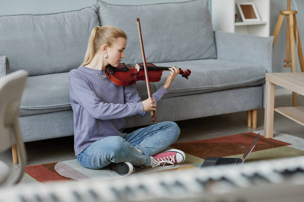 Plná délka portrét blondýny mladé ženy hraje doma housle, zatímco sedí na podlaze v útulném interiéru, kopírovací prostor - Fotografie, Obrázek