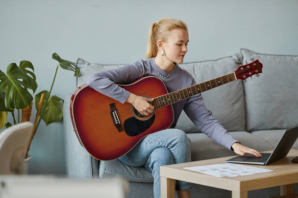 Retrato mínimo de una joven rubia tocando la guitarra en casa y componiendo música mientras está sentada en el sofá, espacio para copiar - Foto, imagen