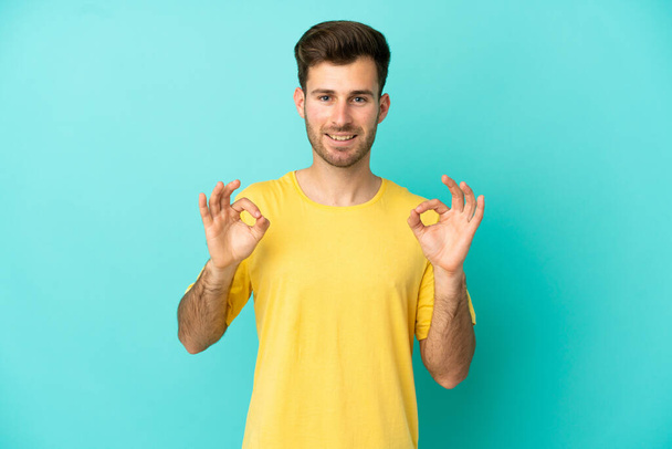 Молодой белый красивый мужчина на голубом фоне показывает знак ОК с двумя руками - Фото, изображение
