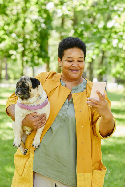 Retrato vertical de una mujer negra madura tomando foto selfie con un lindo perro en el parque y sonriendo felizmente - Foto, Imagen