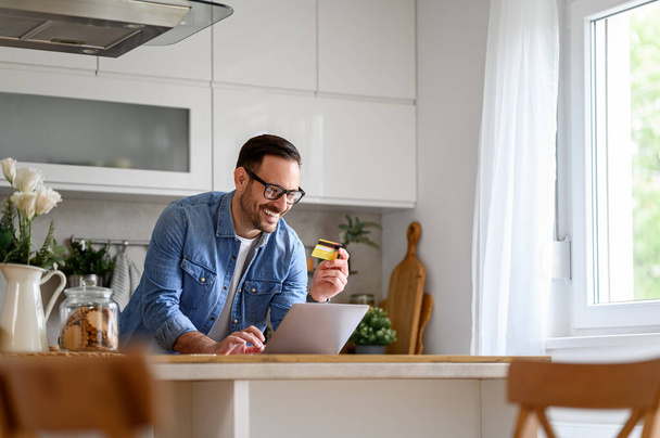 Freelancer masculino feliz añadiendo los detalles de la tarjeta de crédito en el ordenador portátil mientras que compras en línea en el mostrador de cocina - Foto, imagen
