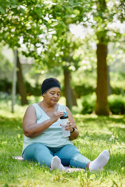 Ritratto verticale a figura intera di donna nera sovrappeso che lavora all'aperto e beve acqua per mantenere l'idratazione - Foto, immagini
