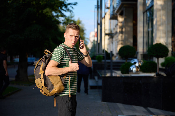 Mladý sportovní muž v pruhovaném tričku kráčející po ulici s chytrým telefonem v ruce a módním hipsterským batohem přehozeným přes rameno a užívající si klidný městský výlet. - Fotografie, Obrázek