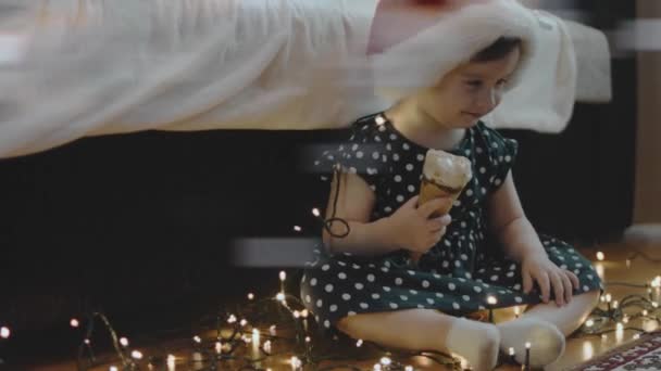 Мила маленька дівчинка в капелюсі Сантаса їсть морозиво на Різдвяній вечірці. Різдвяні вогні навколо - Кадри, відео