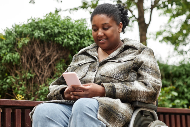 Portret dorosłej czarnej kobiety na wózku inwalidzkim korzystającej ze smartfona na zewnątrz i uśmiechającej się - Zdjęcie, obraz