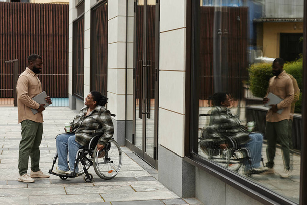 Colpo grandangolare di donna nera in sedia a rotelle che parla con un amico o un collega all'aperto in ambiente urbano, copia lo spazio - Foto, immagini