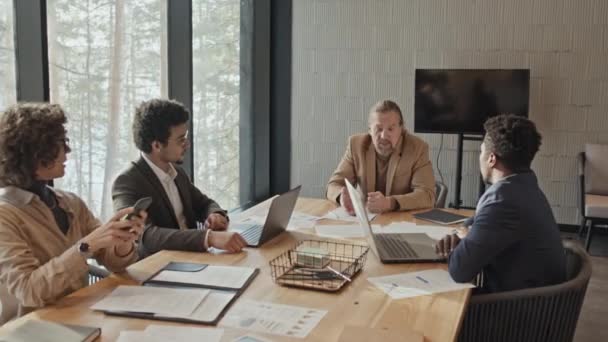 Tiro médio de executivos caucasianos de meia-idade desapontados conversando com a equipe de projeto multiétnico na sala de reuniões no escritório, com laptops, impressões de apresentação e documentos na mesa - Filmagem, Vídeo
