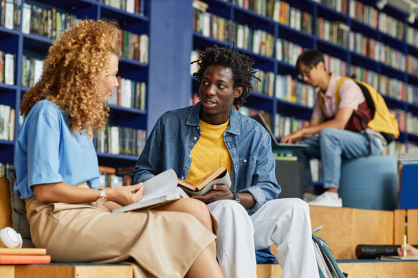 Разнообразная группа студентов, занимающихся в библиотечном зале, акцент на черном молодом человеке, разговаривающем с подругой - Фото, изображение
