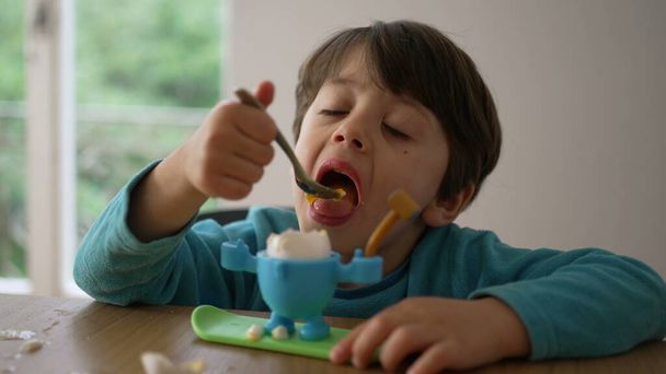 Bambino che mangia uova soffici a colazione con cucchiaio. Ragazzino di 4 anni che mangia da solo, faccia da primo piano assorbita in uno spuntino sano, oeuf a la coque - Foto, immagini
