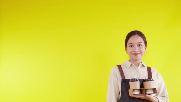Retrato joven barista asiática mujer usando delantal sosteniendo taza de café y presentando sobre fondo amarillo, camarera o emprendedor alegre y mostrando y pulgares hacia arriba, pequeña empresa o puesta en marcha. - Metraje, vídeo