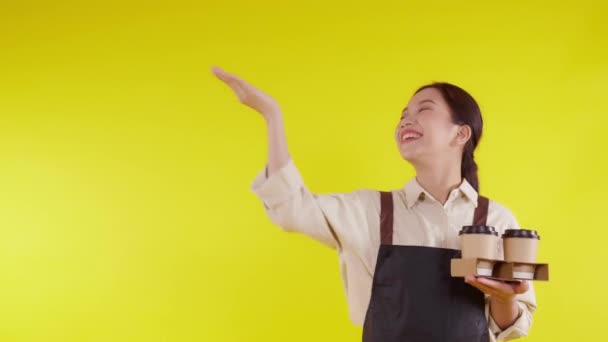 Retrato joven barista asiática mujer usando delantal sosteniendo taza de café y presentando sobre fondo amarillo, camarera o emprendedor alegre y mostrando y pulgares hacia arriba, pequeña empresa o puesta en marcha. - Metraje, vídeo