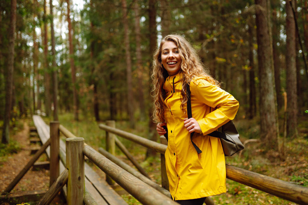 Woman traveler in a bright coat walks along a wooden path among the wetlands. A tourist with a backpack walks through the forest. Travel, nature concept. - Fotoğraf, Görsel