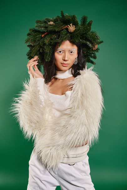 winter concept, asian woman with white makeup and trendy outfit posing in wreath on green backdrop - Photo, Image