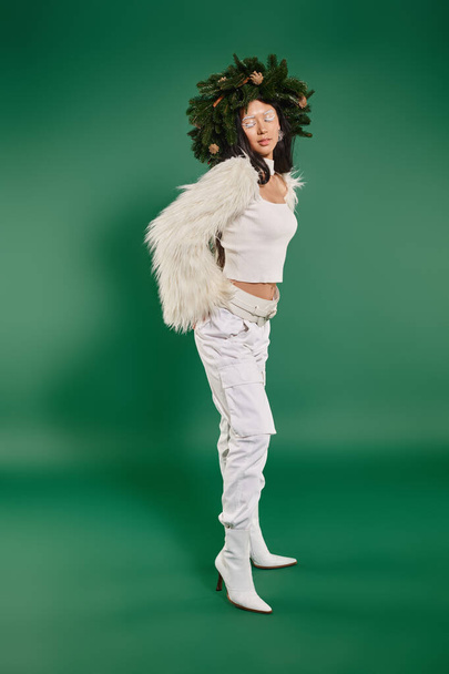 εορταστική εποχή, Ασιάτισσα γυναίκα με λευκό μακιγιάζ και μοντέρνα εμφάνιση σε στεφάνι σε πράσινο φόντο - Φωτογραφία, εικόνα