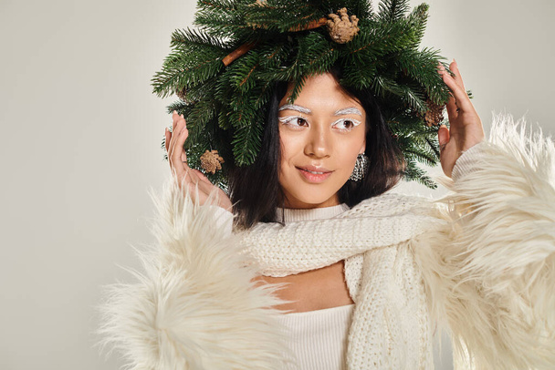 winter schoonheid, positieve vrouw met natuurlijke pijnboom krans poseren in witte kleren op grijze achtergrond - Foto, afbeelding