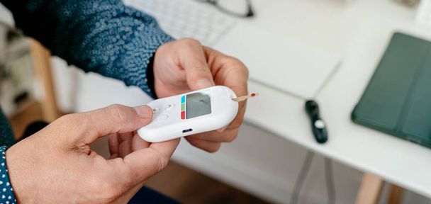 un homme caucasien mesure son taux de glucose sanguin en plaçant une bandelette dans un glucomètre au bureau, dans un format panoramique à utiliser comme bannière Web ou en-tête - Photo, image