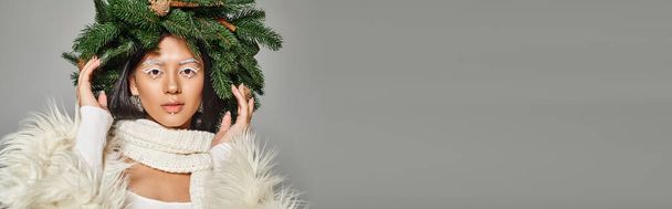winter banner, attractive woman with white eye makeup and beads on face posing in wreath on grey - Photo, Image