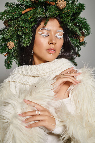 holiday style, attractive woman with white eye makeup and beads on face posing in wreath on grey - Photo, Image