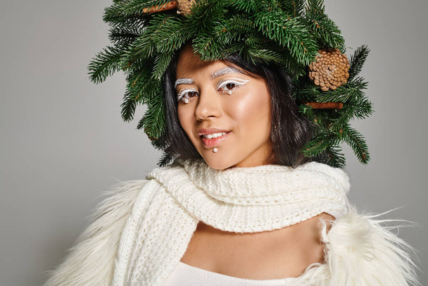 happy woman with white eye makeup and beads on face posing in wreath on grey backdrop, winter queen - Photo, Image