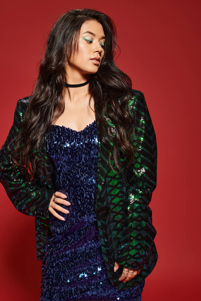 brunette asian woman in green dress and jacket with sequins posing with hand on hip on red backdrop - Photo, Image