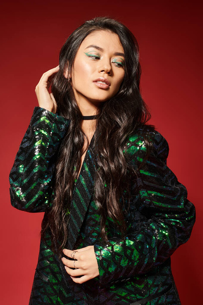 asian brunette woman in trendy green jacket with sequins adjusting wavy hair on red background - Foto, immagini