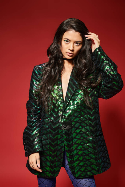 beautiful asian woman in trendy green jacket with sequins adjusting wavy hair on red background - Foto, Imagem
