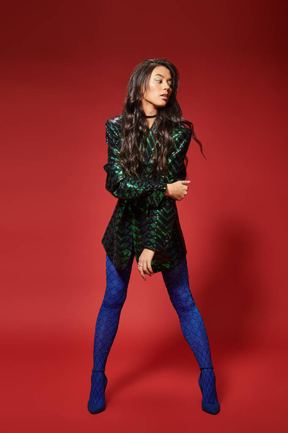 attractive woman in trendy green jacket with sequins and blue pantyhose posing on red background - Foto, Imagem