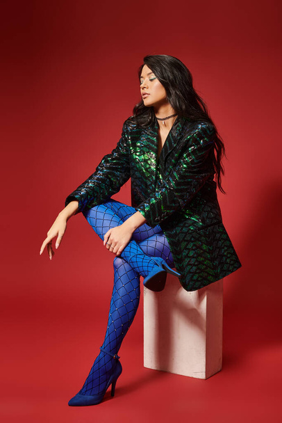 trendy asian woman in green jacket with sequins and blue pantyhose sitting on red backdrop - Foto, immagini