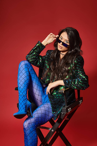 stylish asian woman in sunglasses and green jacket with sequins sitting on chair on red backdrop - Photo, Image