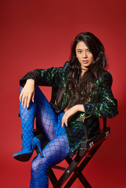 stylish asian woman in green jacket with sequins and blue pantyhose sitting on chair on red backdrop - Foto, immagini