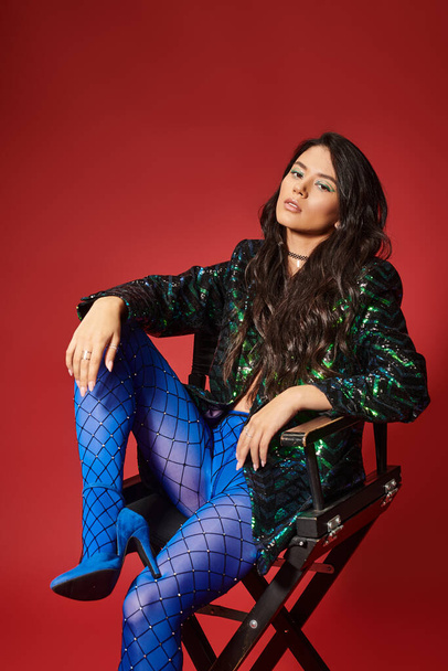 pretty asian woman in green jacket with sequins and blue pantyhose sitting on chair on red backdrop - Foto, immagini