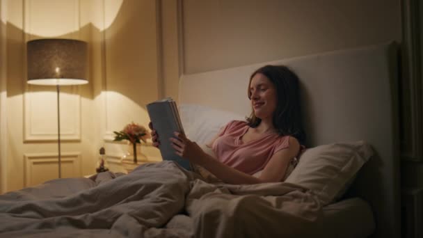 Friedliche Frau liest Buch am Abend zu Hause. Lächelnder Leser, der in der Wochenendnacht in seinem gemütlichen Bett die neuartige Ruhe genießt. Ruhige, entspannte Weibchen, die spät im Lampenlicht lernen. Lieblingsliteratur Hobbykonzept. - Filmmaterial, Video