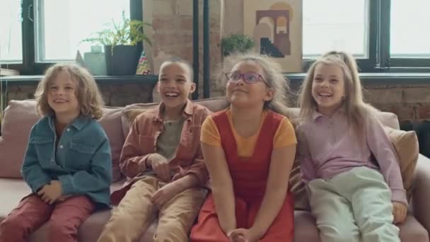 Mittlere Aufnahme einer Gruppe von vier verschiedenen Jungen und Mädchen in lässiger Kleidung, die zusammen auf der Couch zu Hause sitzen und lustige Darbietungen oder Shows sehen, lächeln und lachen - Filmmaterial, Video