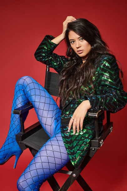 brunette asian woman in trendy jacket with sequins and blue pantyhose sitting on chair, red backdrop - Valokuva, kuva