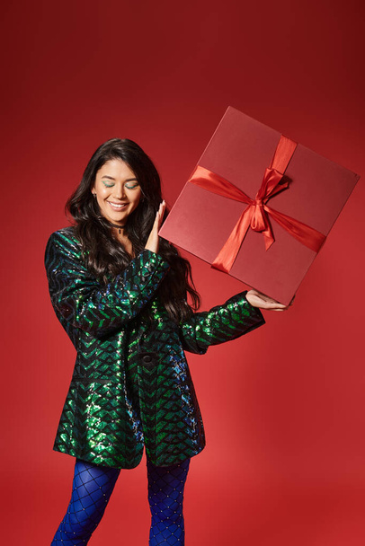 happy asian woman in jacket with sequins and blue pantyhose holding present on red backdrop - 写真・画像