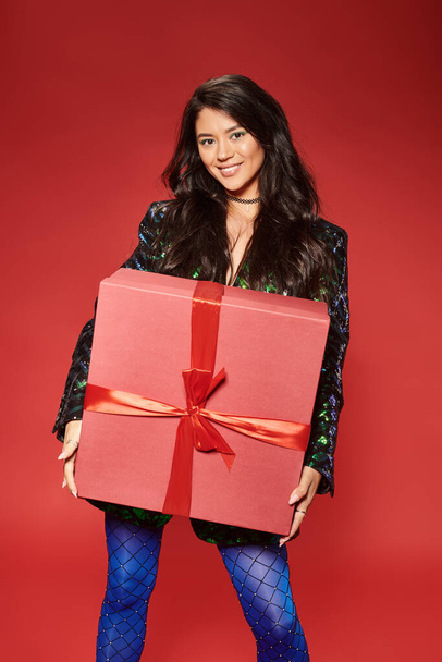 χαρούμενη Ασιάτισσα με πράσινο σακάκι με πούλιες που κρατούν κουτί δώρου σε κόκκινο φόντο, Καλά Χριστούγεννα - Φωτογραφία, εικόνα