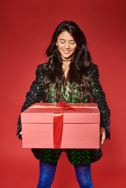 alegre asiático mulher no verde jaqueta com lantejoulas olhando para presente caixa no vermelho pano de fundo, Feliz Natal - Foto, Imagem