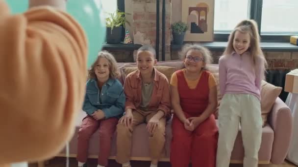 Середній повноплемінний знімок групи дев'ятирічних багатоетнічних дітей, які сидять на дивані на вечірці на день народження і спостерігають за невпізнаваним артистом в костюмах виконують трюки з повітряною кулею - Кадри, відео