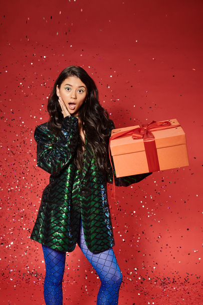 Buon Natale, donna asiatica scioccata in giacca verde con paillettes regalo di apertura, coriandoli festivi - Foto, immagini