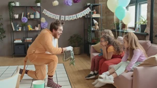 Full shot de animador masculino alegre en traje de gato y maquillaje demostrando trucos de fiesta con manzana y tela para el grupo de niños preadolescentes sentados en el sofá, que están viendo con sorpresa - Metraje, vídeo