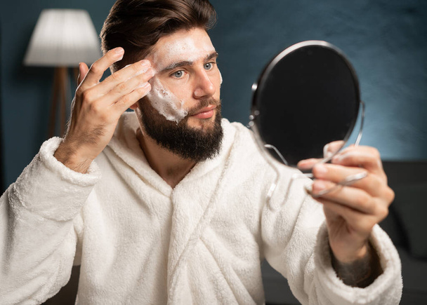 Hombre guapo aplicando mascarilla de sábana en su cara y mirándose en el espejo en casa. Primer plano - Foto, imagen