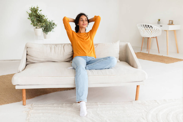 Dama árabe casual descansando en el sofá con sonrisa contenta tomando un descanso en su moderna sala de estar, apreciando su tiempo libre en casa. Abrazando un momento de calma doméstica - Foto, imagen