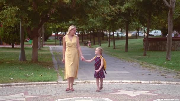 Mutlu anne ve küçük kızı okula gidiyor. Şehir parkında yürüyorlar. Okul kızı, çocuğuyla birlikte şehir bahçesinde ileriyi gösteren kızın elini tutuyor. - Video, Çekim