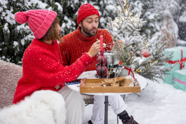 Man and woman in red winter clothes have fun while eating fondue, sitting together by the table outdoors at snowy garden. Young family celebrating winter holidays - Photo, image